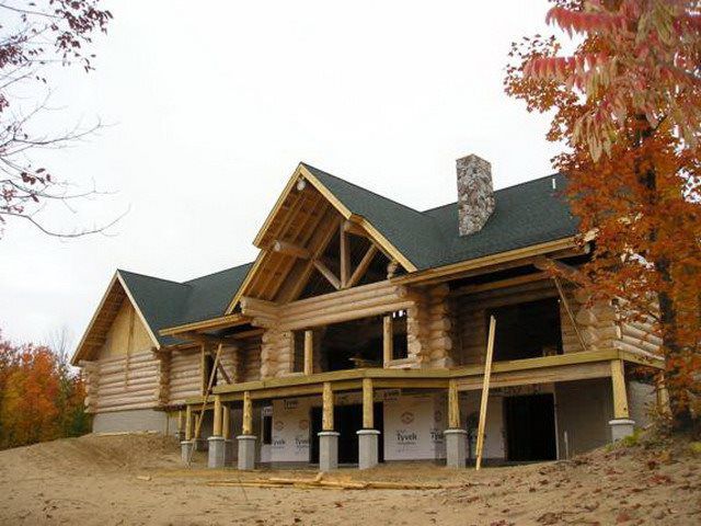 Log cabin under construction with exposed beams, green roof, and stone chimney. Wooden deck. 