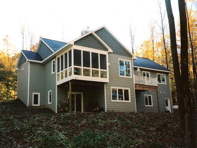 Two-story house with screened porch and deck, green siding, set in wooded area.