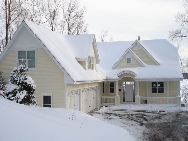 Yellow house with snow-covered roof and surroundings, three-car garage, porch, and a wintery landscape.
