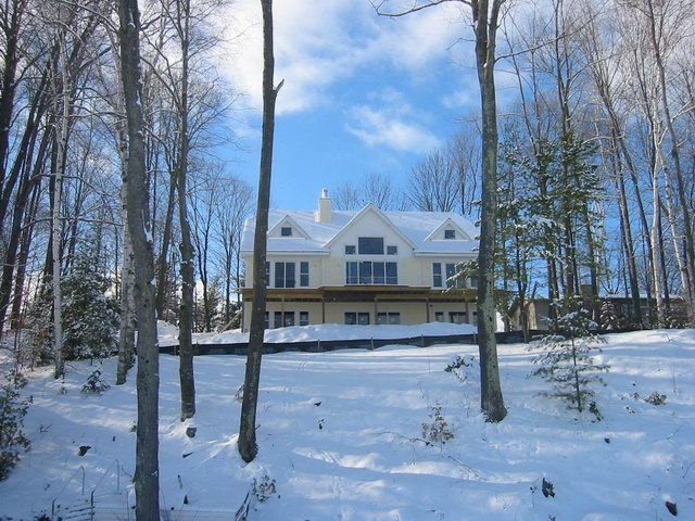 Snow-covered hillside with trees framing a two-story beige house under a blue sky.