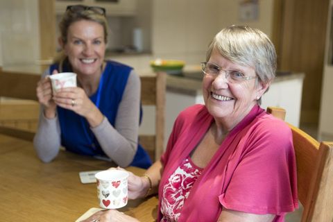 Community care worker joins an an elderly woman for a cup of tea while sitting at her kitchen table