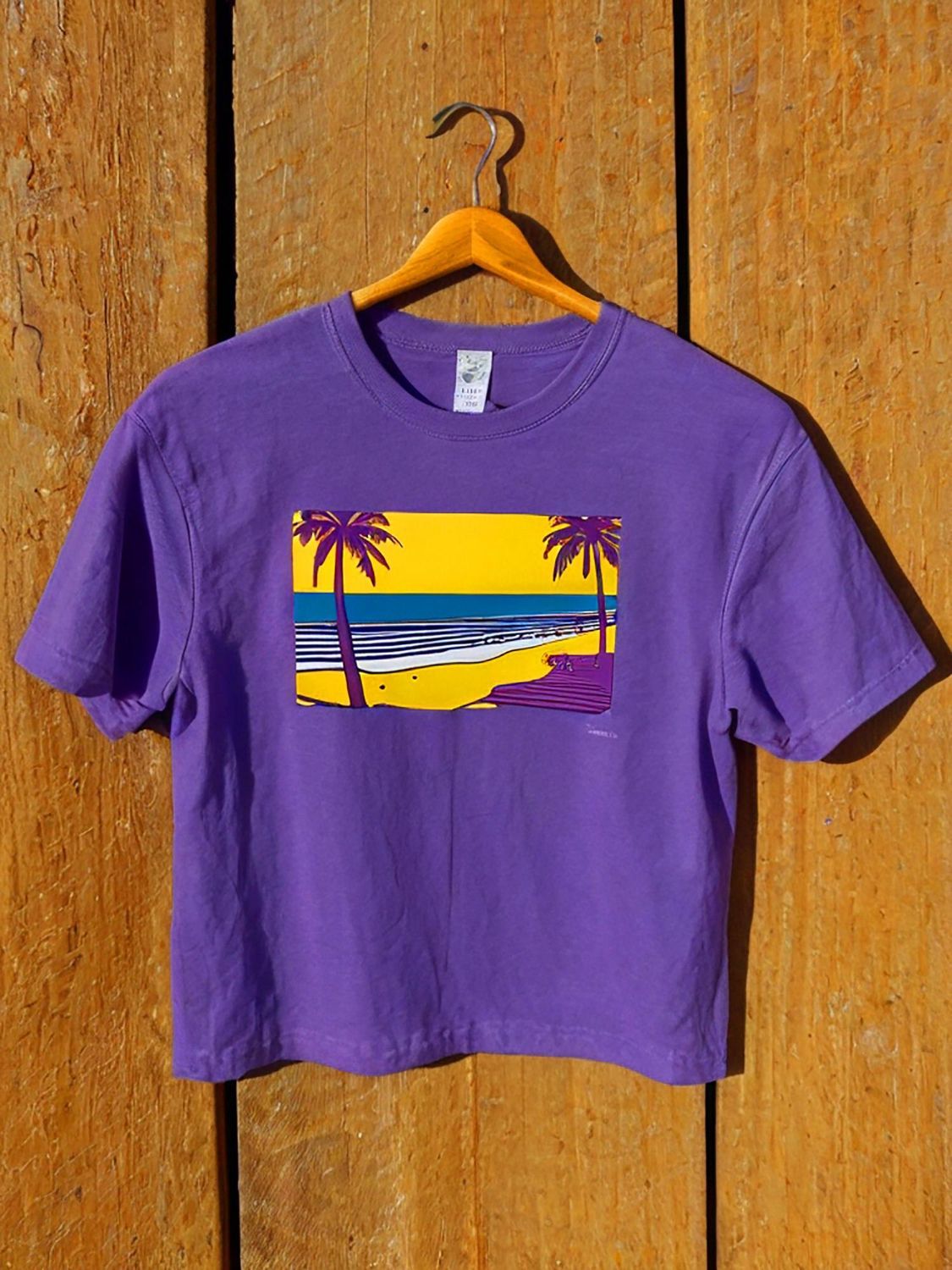 Purple t-shirt with a DTF (Direct To Film) printed beach scene: yellow sky, blue water, palm trees, and purple shore.