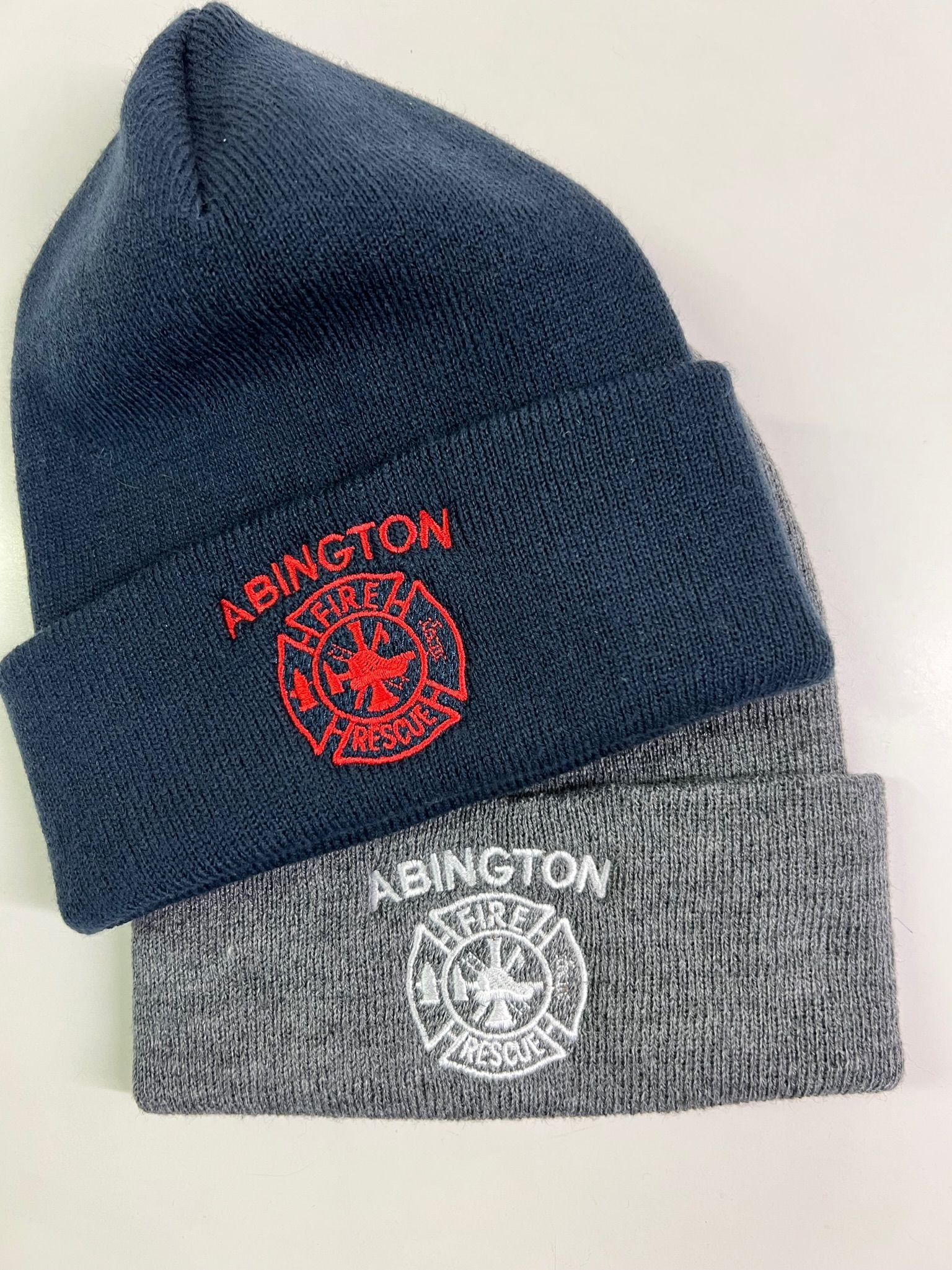 Two knitted beanies, one dark blue with red embroidery, one gray with white embroidery. Both have 
