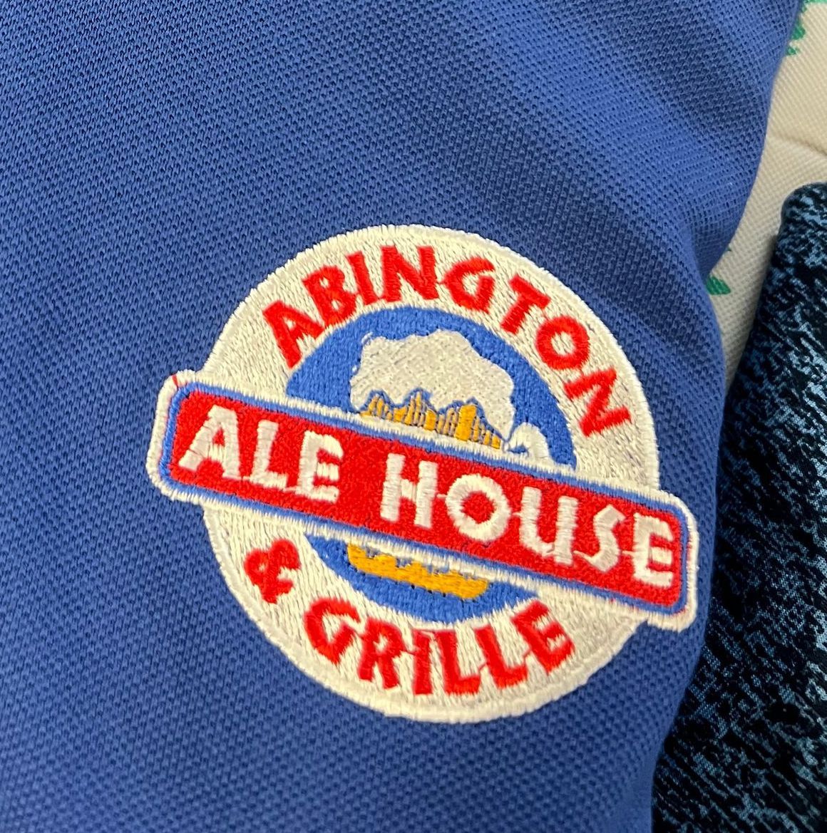Custom embroidery featuring Abington Ale House & Grille logo patch on a blue shirt. Round logo with beer illustration and red text.