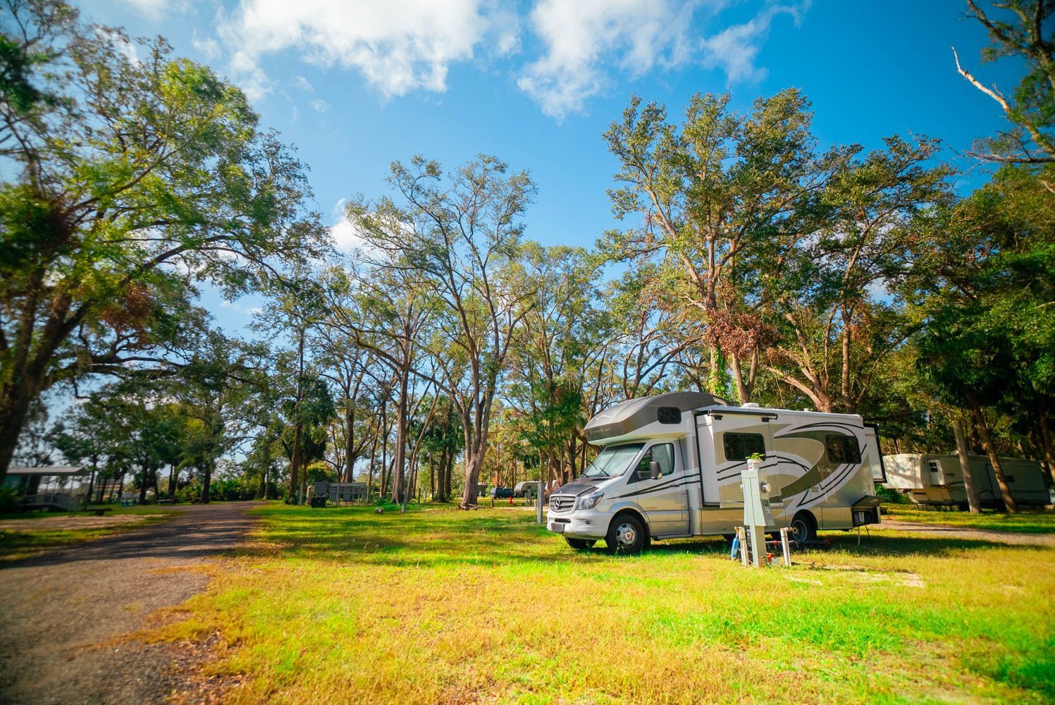 A rv is parked in a grassy field with a guitar , chairs and a surfboard.