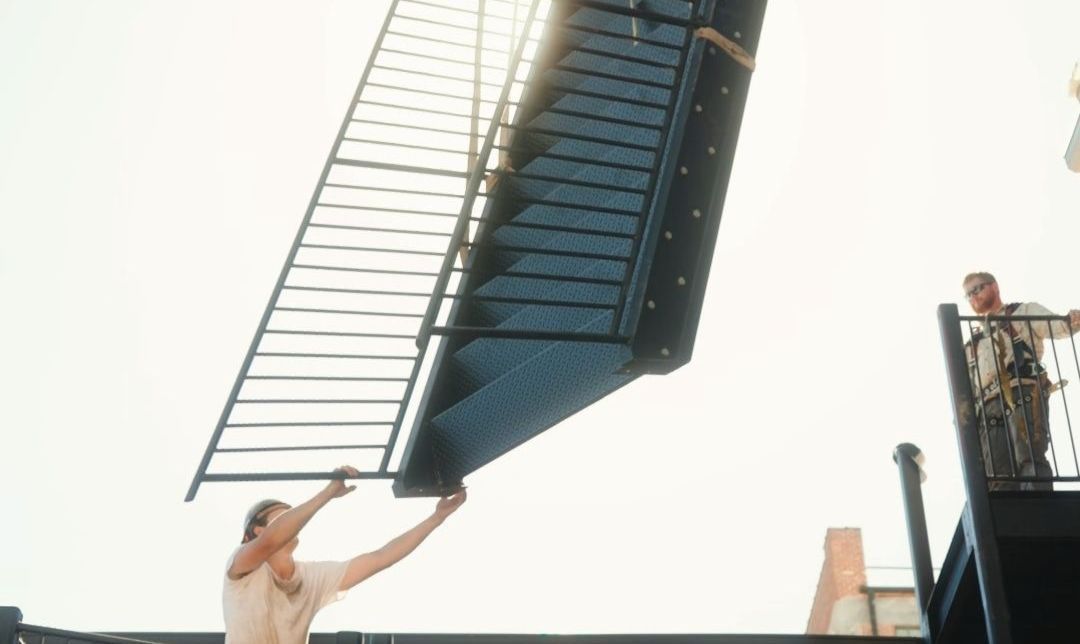 Man holding black staircase, another man watches from above. Bright sky.