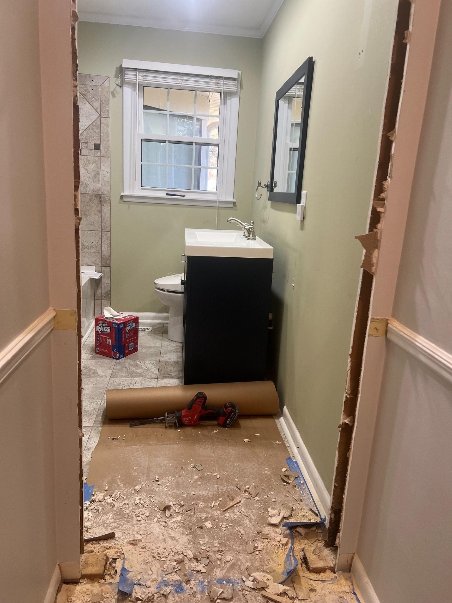 Bathroom renovation in progress, doorway view. Green walls, black vanity, debris on floor, door frames removed.