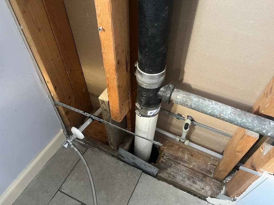 Plumbing pipes exposed in a wall. Black drain pipe, grey vent pipe, wood framing, and tile floor.