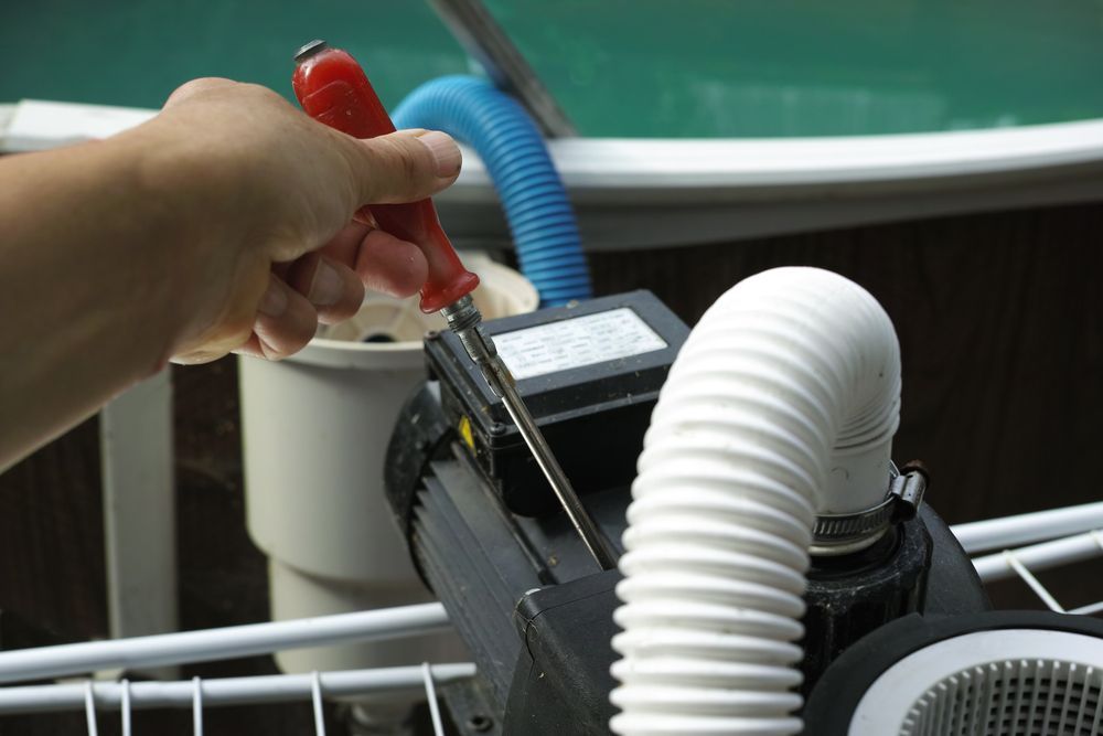 Hand using screwdriver to adjust pool pump components. Blue and white hoses.