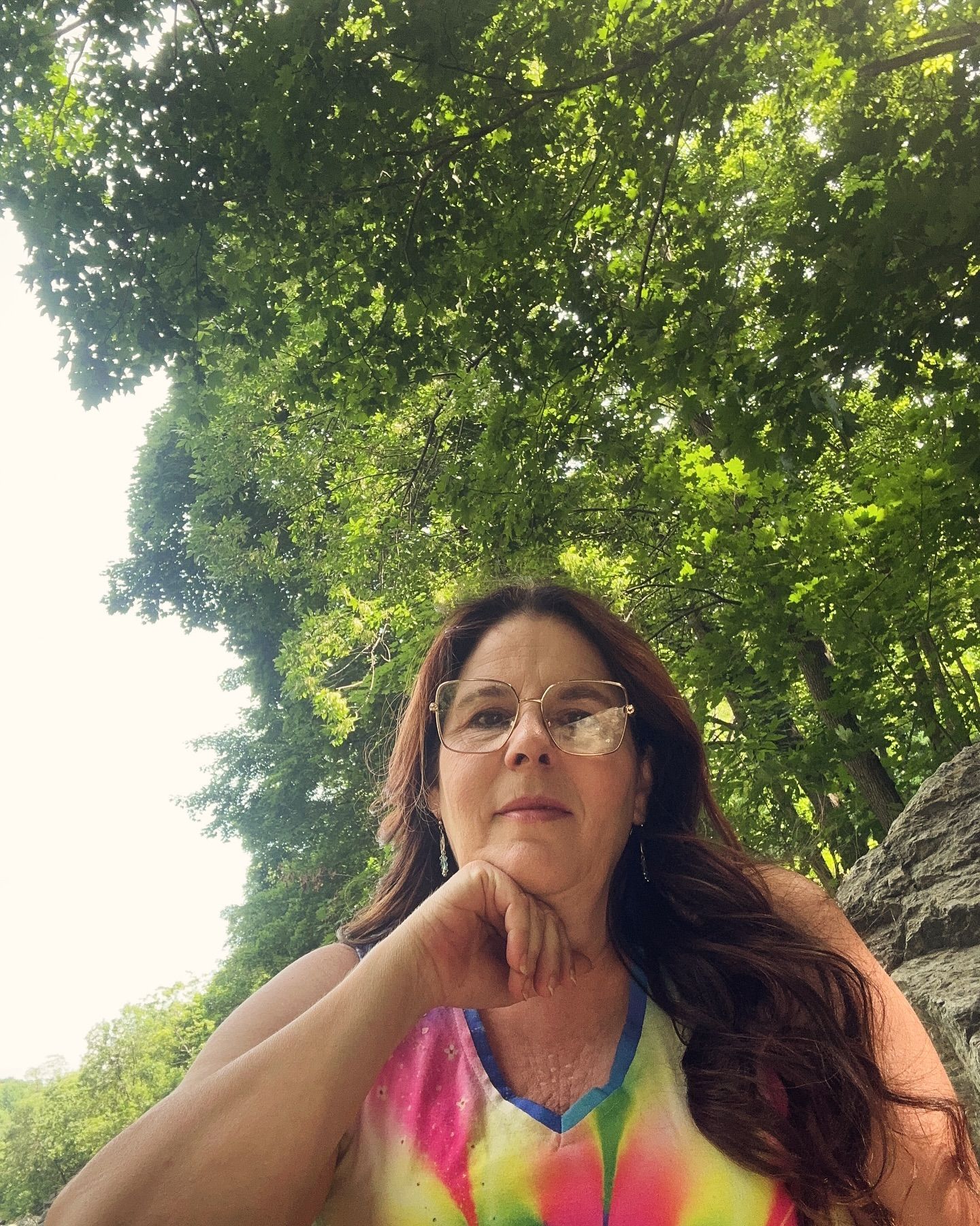 A woman wearing glasses and a tie dye shirt is standing in the woods.
