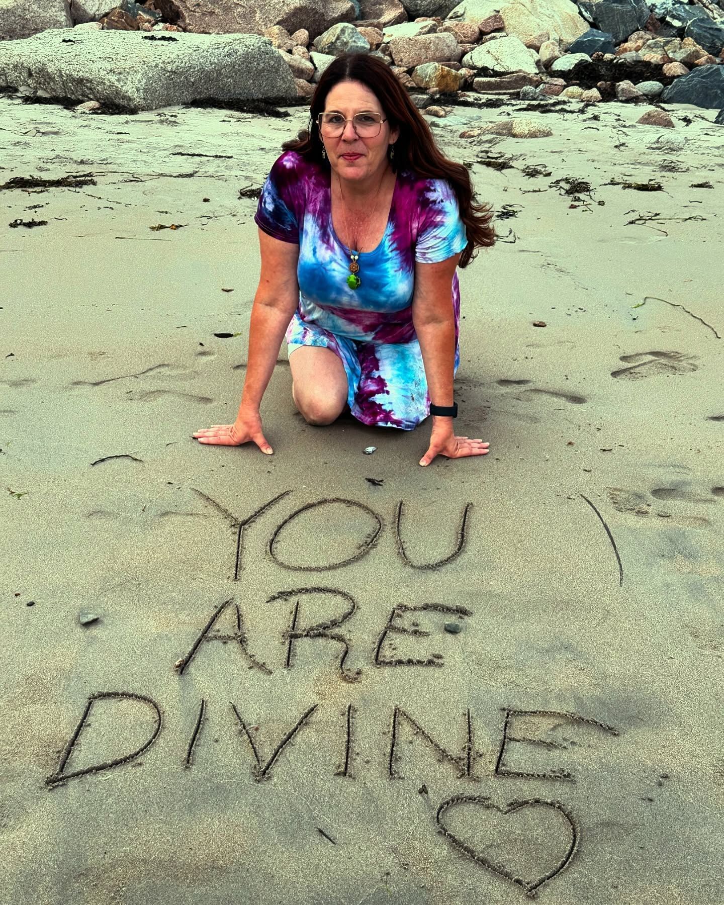 A woman kneeling on the beach with the words you are divine written in the sand