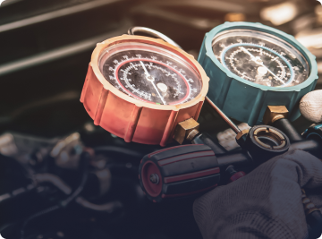 A gloved hand holds an automotive AC manifold gauge set with red and teal gauges over a vehicle engine. | Depot Auto Repair