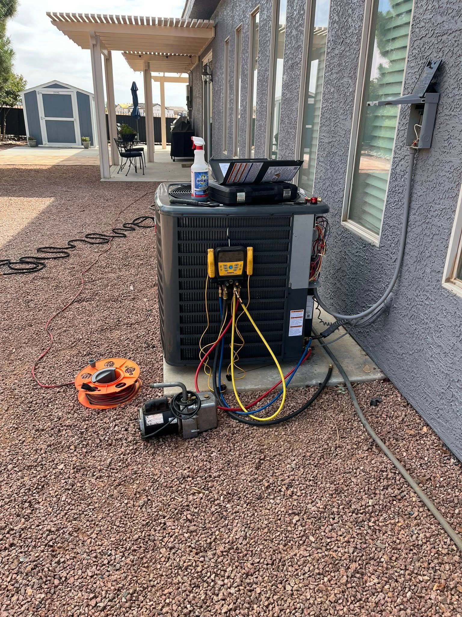 An air conditioning unit outdoors with open wires; tools are placed nearby. The unit sits on gravel next to a house.