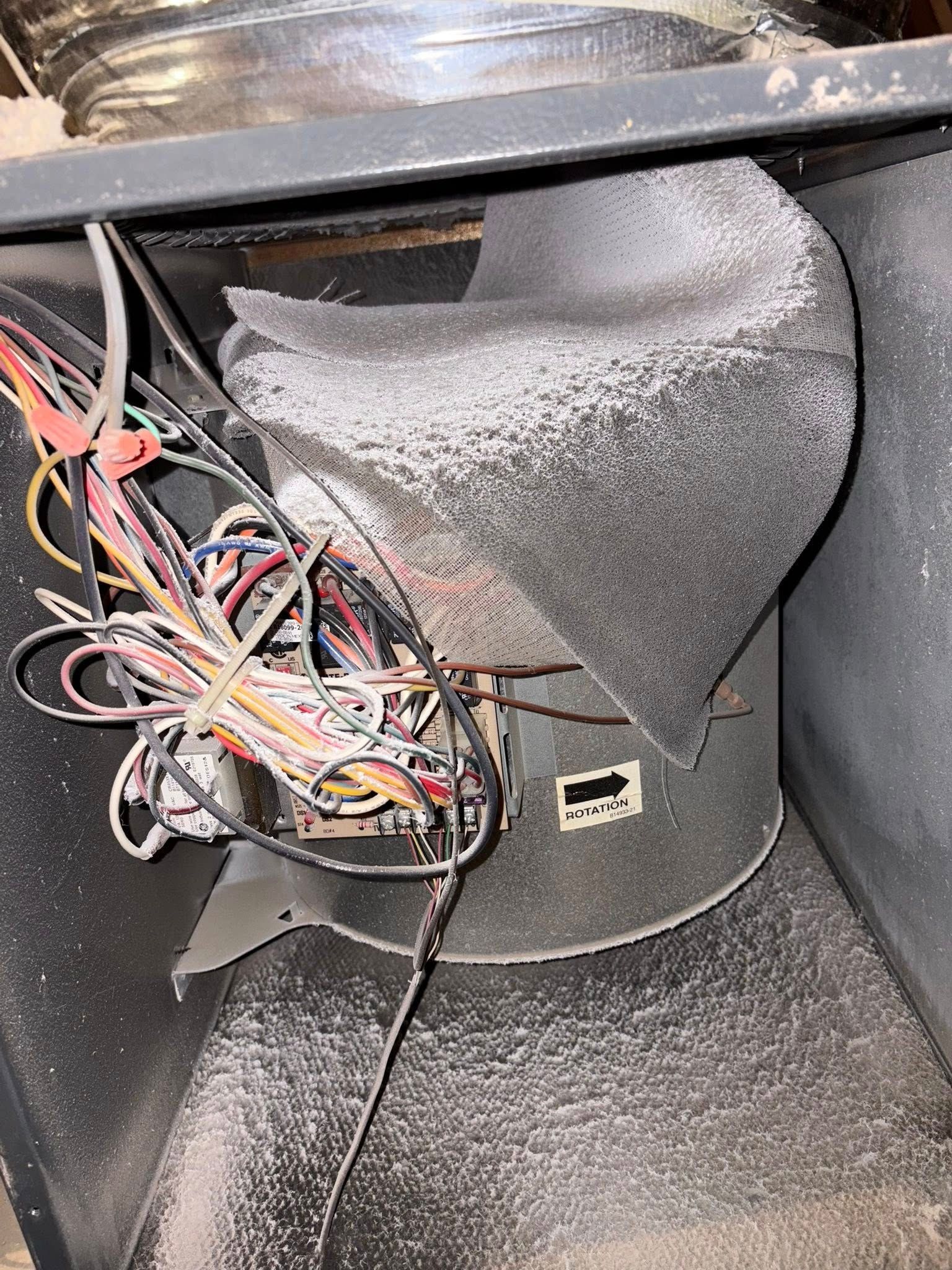 Furnace blower fan with wiring and ductwork, coated in white dust. The fan has an arrow indicating air flow.