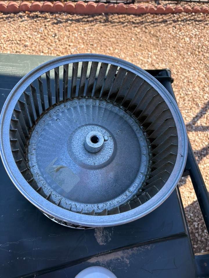 A cylindrical fan blade assembly, likely from an HVAC unit, sits on a dark surface outdoors. Gray metal with many fins.