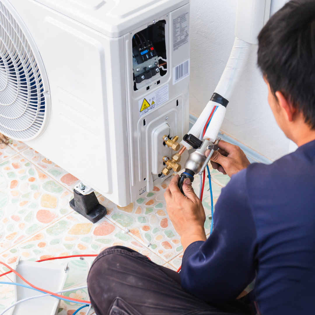 Person in a dark blue shirt works on an air conditioning unit outside. They are connecting tubes and wires.
