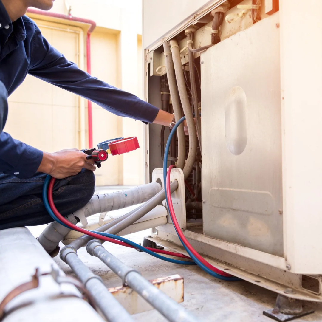 HVAC technician in blue uniform checks an outdoor unit, using gauges and hoses.