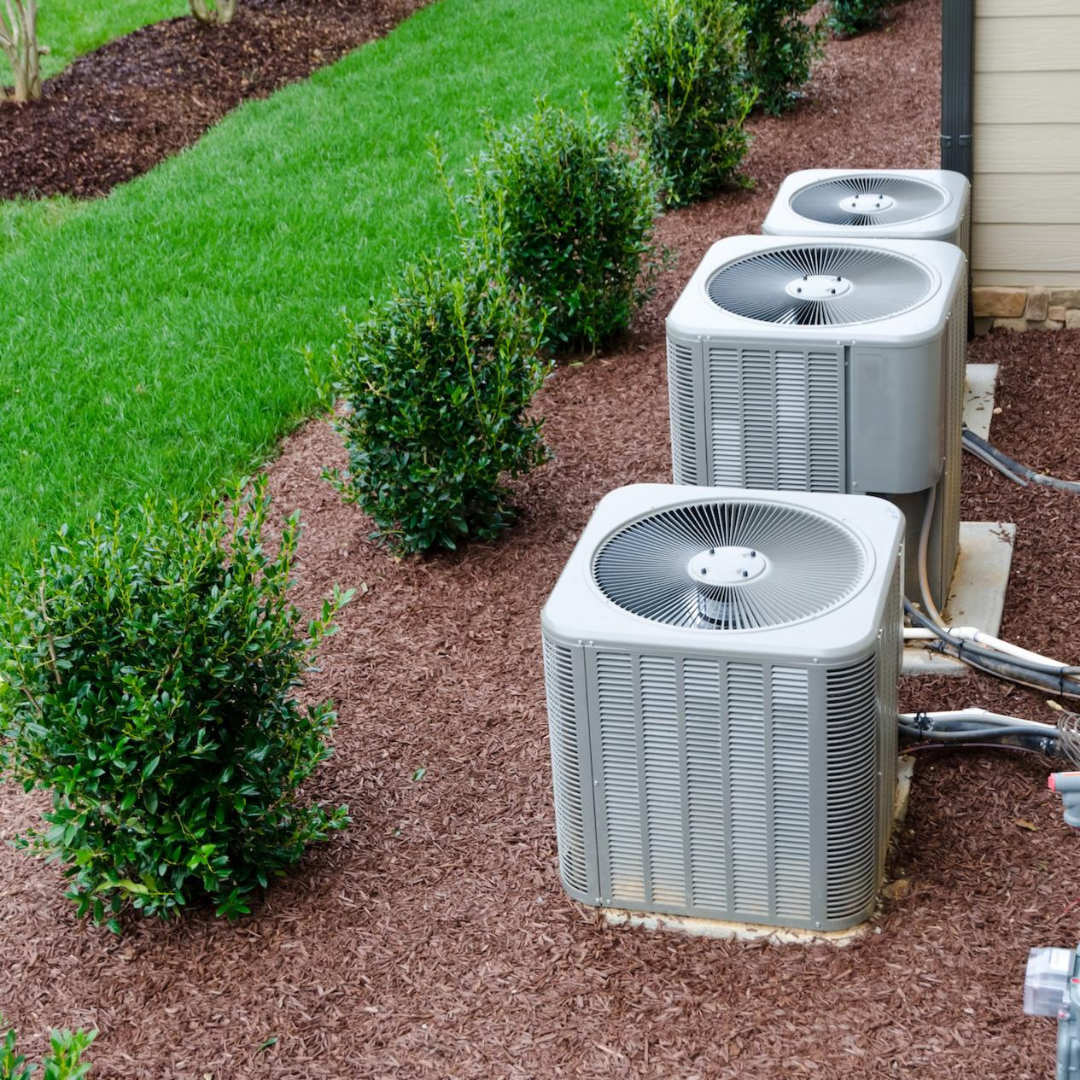 Three air conditioning units in a yard with brown mulch and small green bushes, near a strip of green grass.