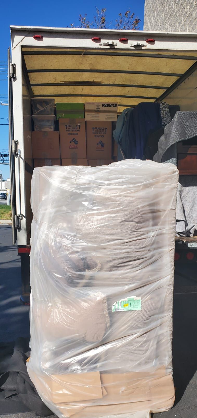 A box truck loaded with items, the foreground has a pallet wrapped in plastic.