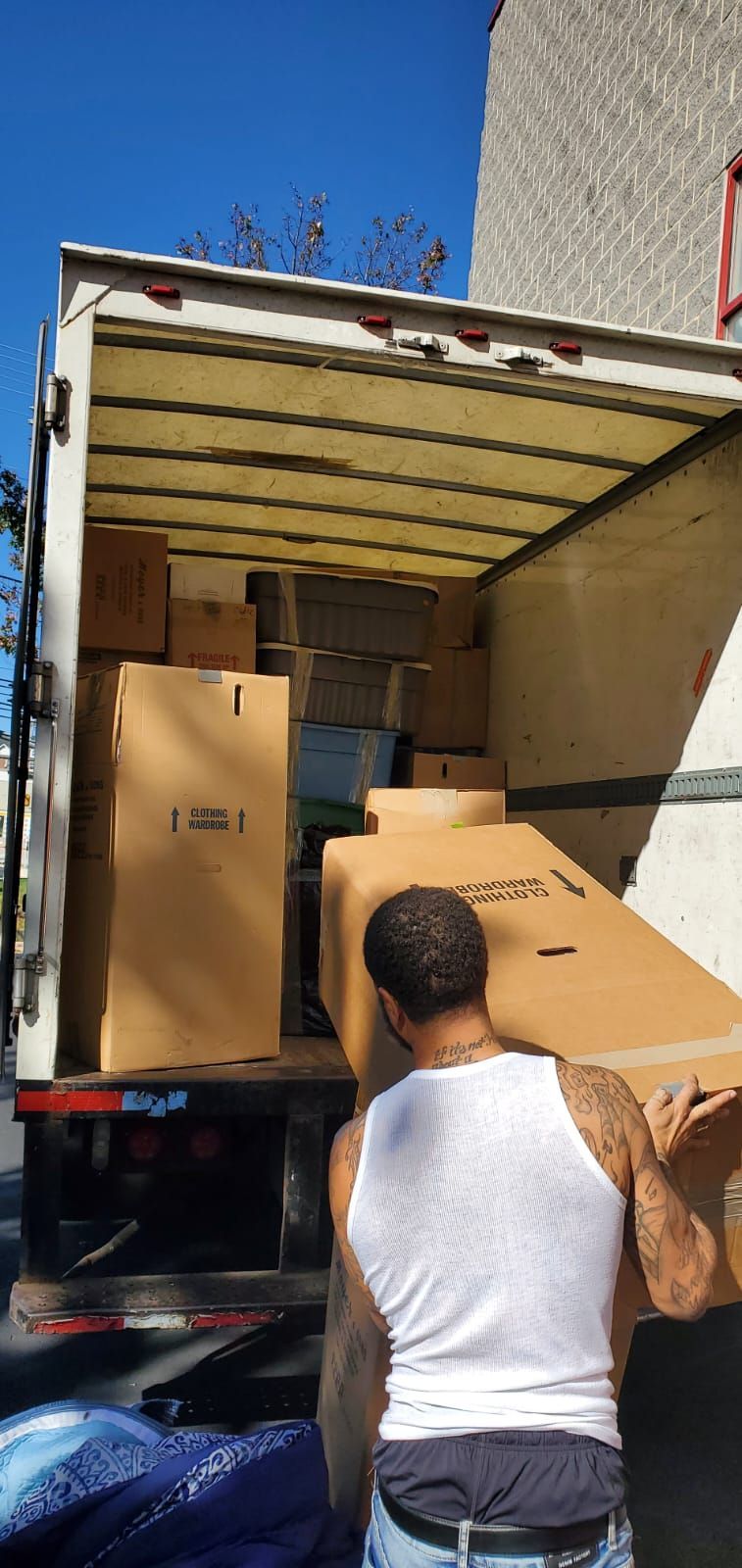 A man in a white tank top loads boxes into a moving truck on a sunny day.