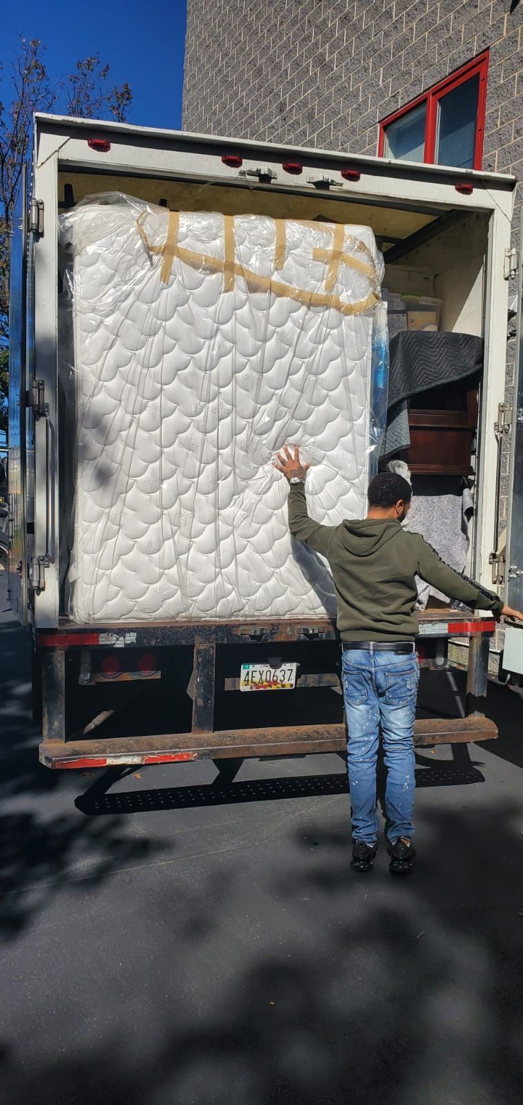 Person standing in front of a truck loaded with a mattress, arms outstretched. Sunny day.