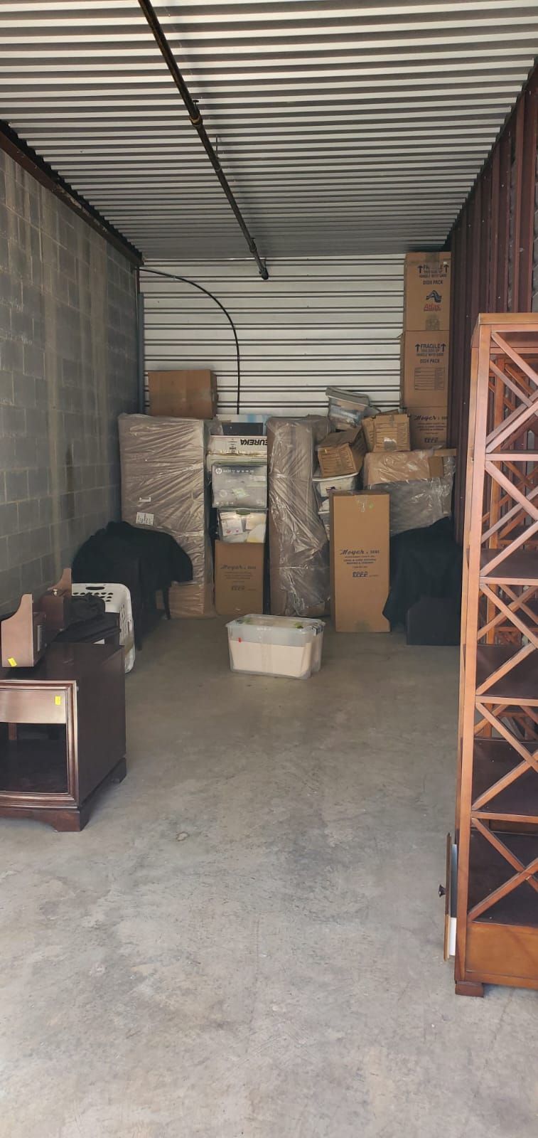 A storage unit with packed boxes, furniture, and a nightstand. The floor and walls are concrete.