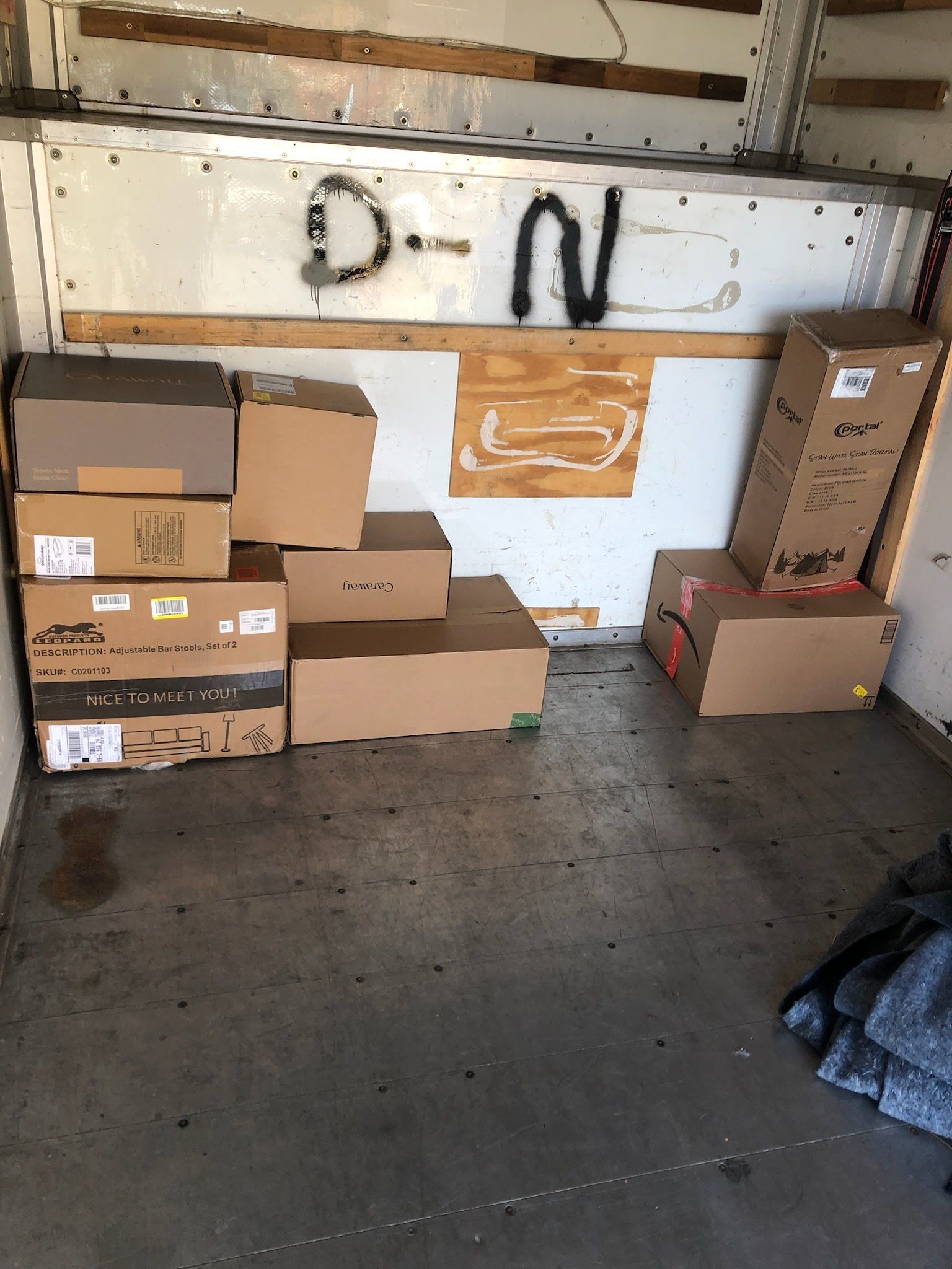 Boxes stacked inside a cargo van. Some cardboard is light brown; the floor is gray; graffiti is visible on the wall.