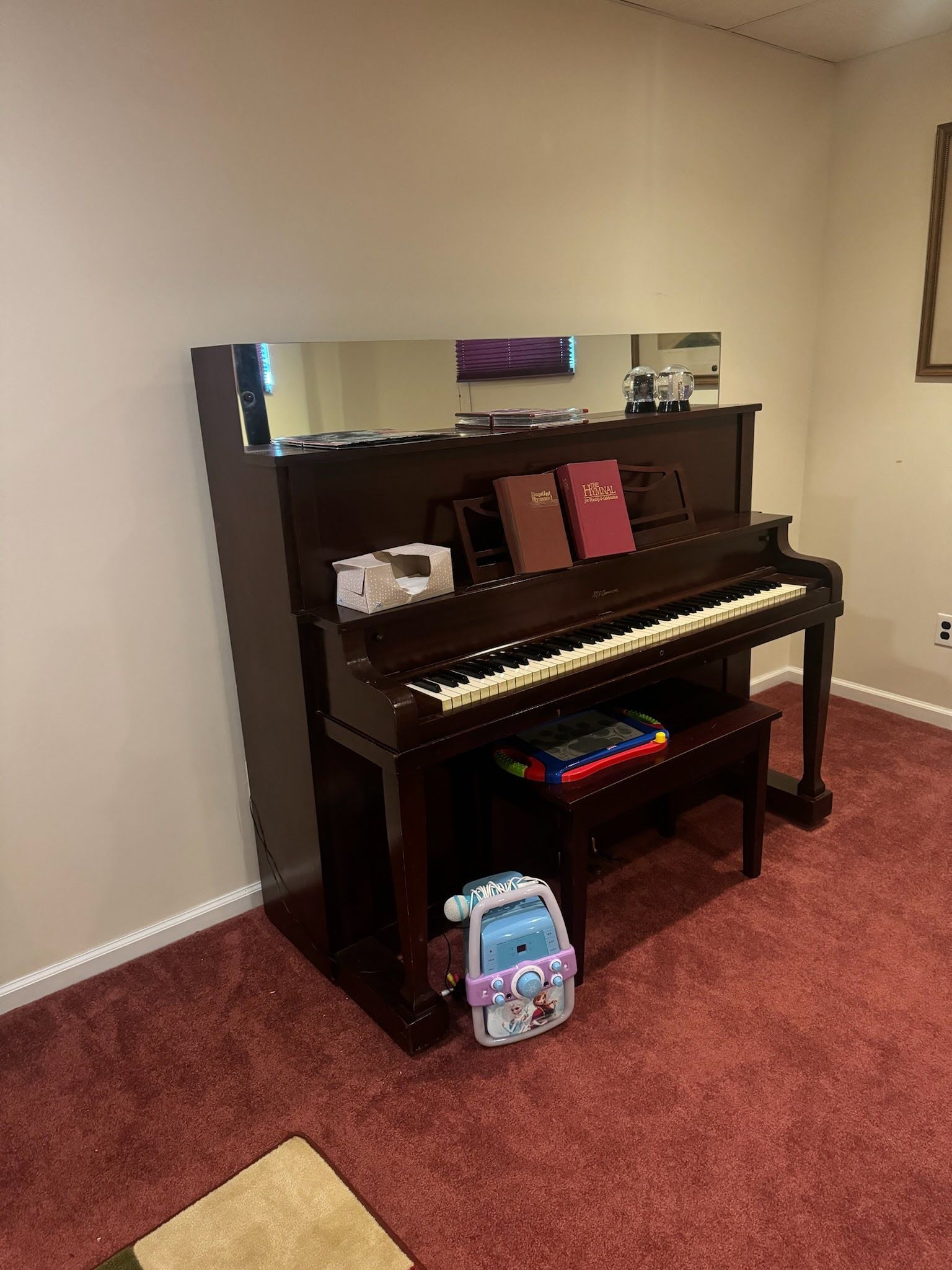Upright piano in a room, with a bench and books, and a child's bag on the floor.
