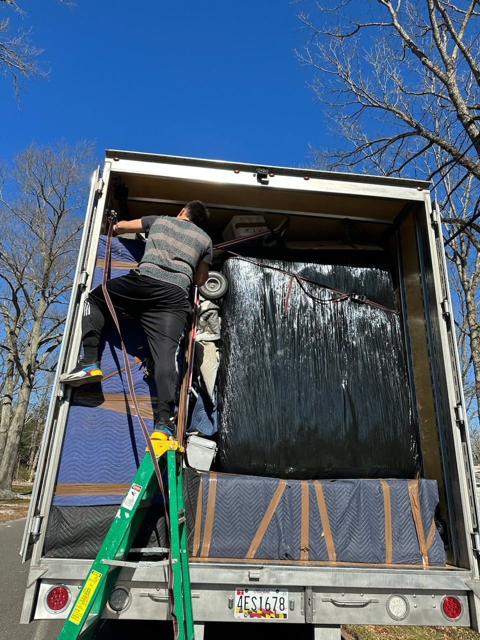 A man on a ladder loads a truck with a large, black-wrapped item.