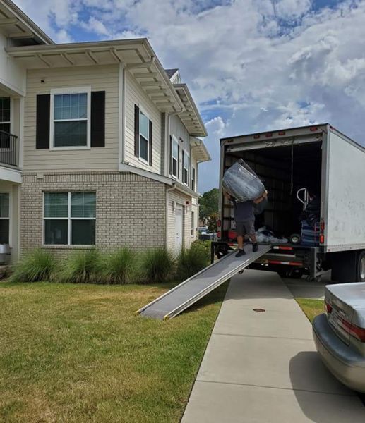 A person loads a moving truck from a building with beige brick, ramp extended, sunny day.