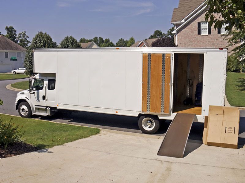 White moving truck parked in front of a house with ramp extended and cardboard boxes nearby.