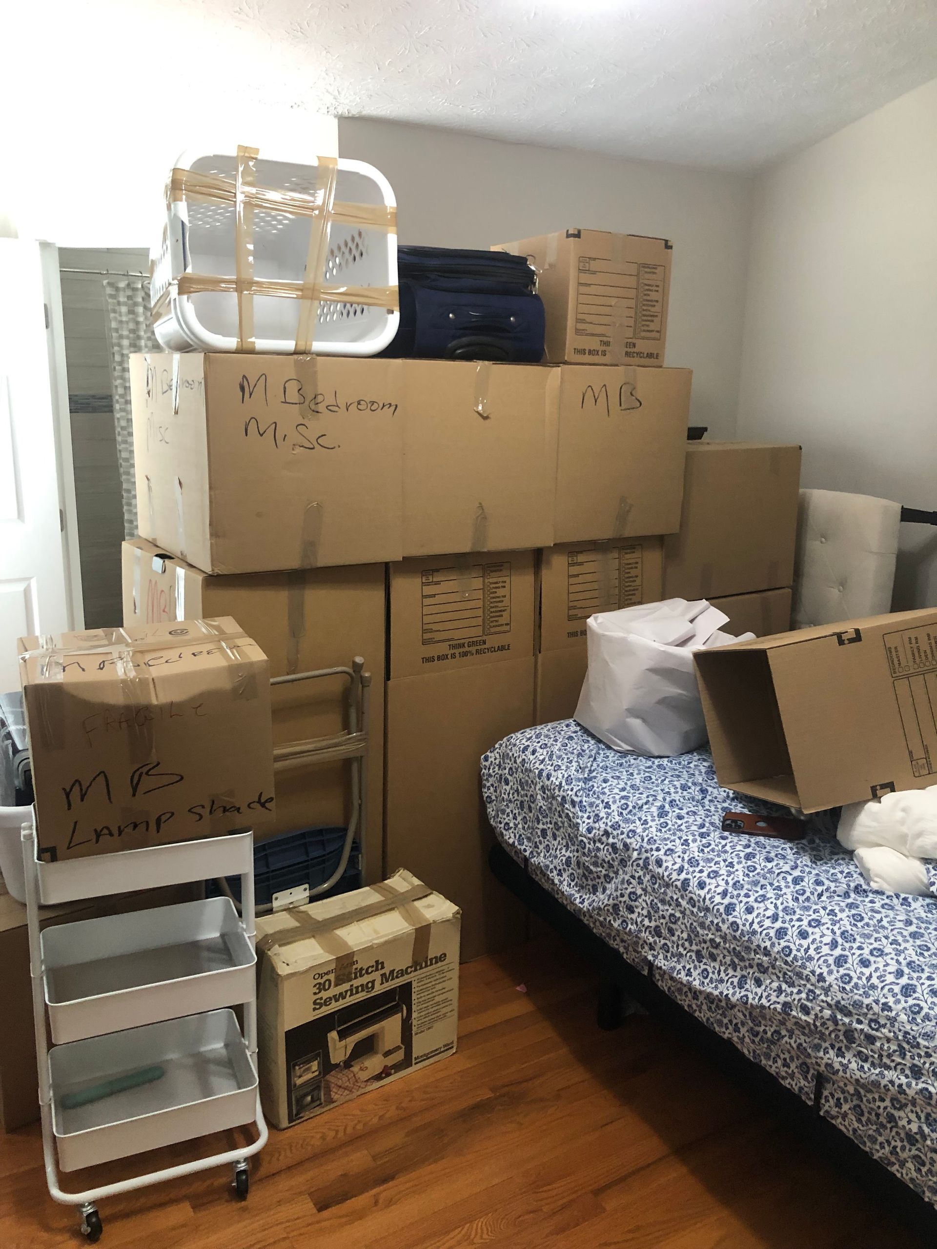 Boxes and belongings stacked in a room, including a bed and rolling cart.