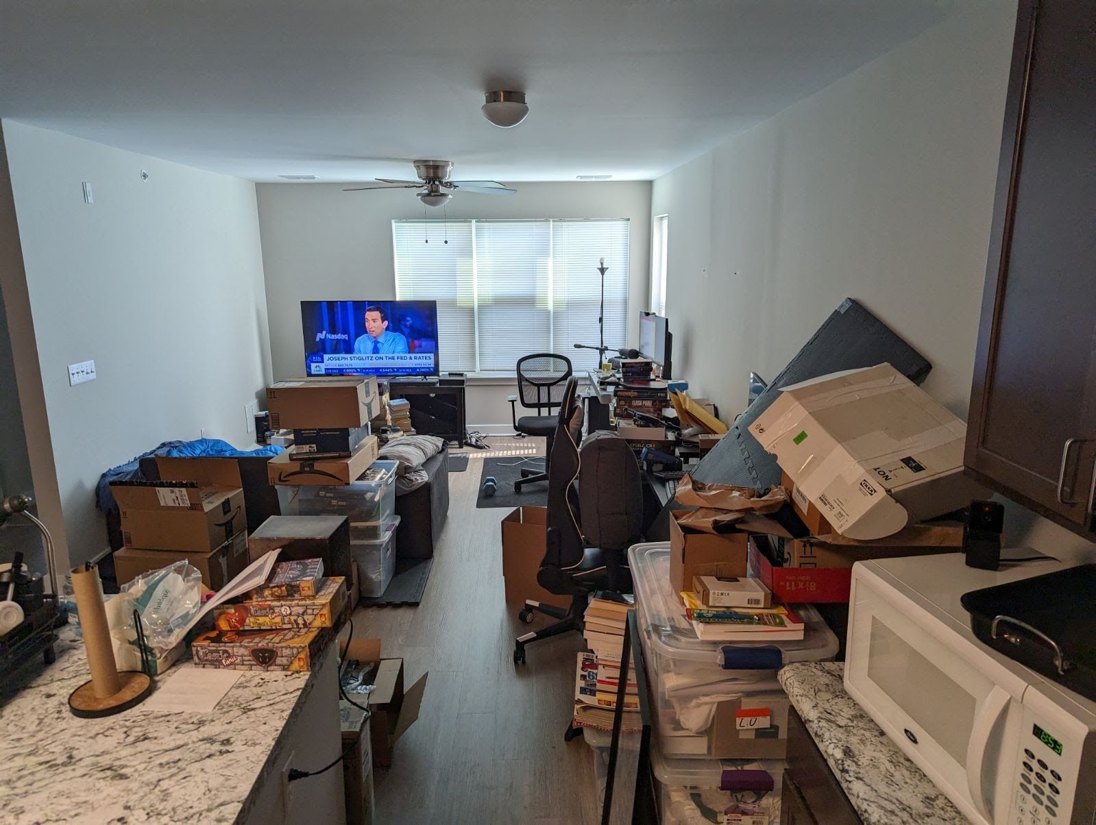 Cluttered living space with boxes, furniture, and a TV; kitchen countertop and microwave in the foreground.