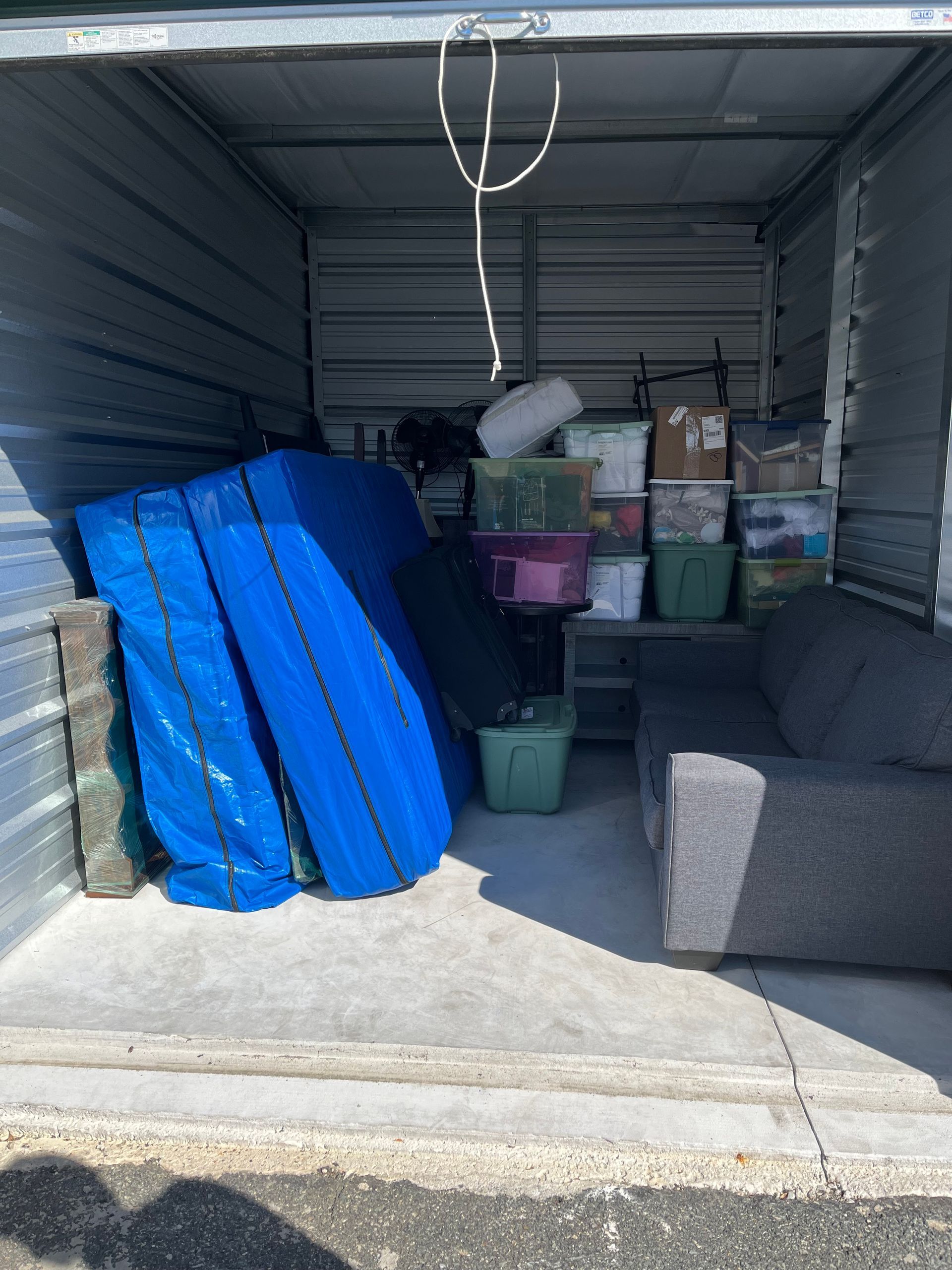 Storage unit with a gray sofa, boxes, blue tarp-covered items, and plastic bins.