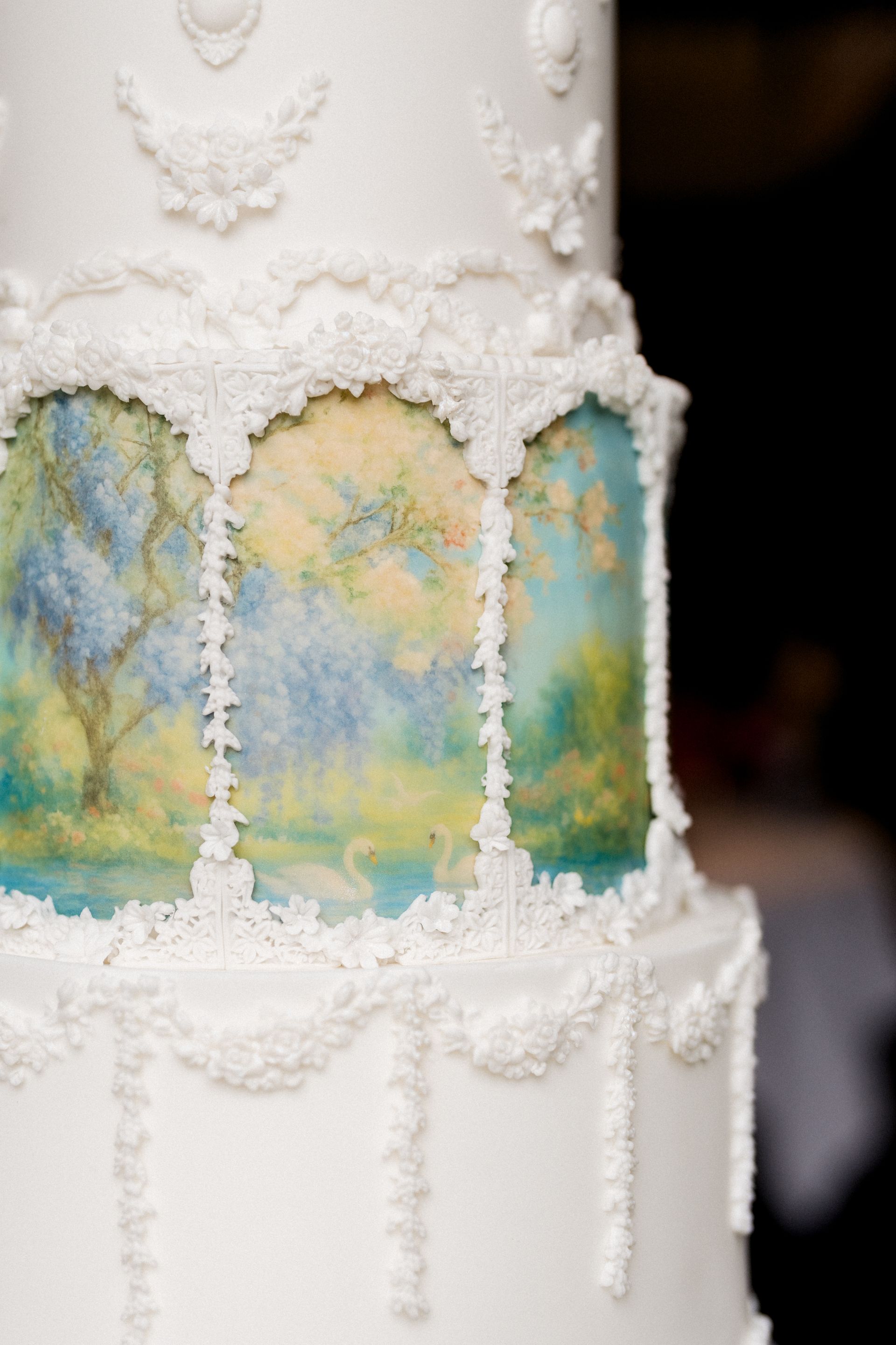 Detailopname van de derde etage van de bruidstaart, met een handgeschilderd Monet-geïnspireerd landschap in blauw-groene tinten, omlijst door witte suiker ornamenten en sierlijke bogen