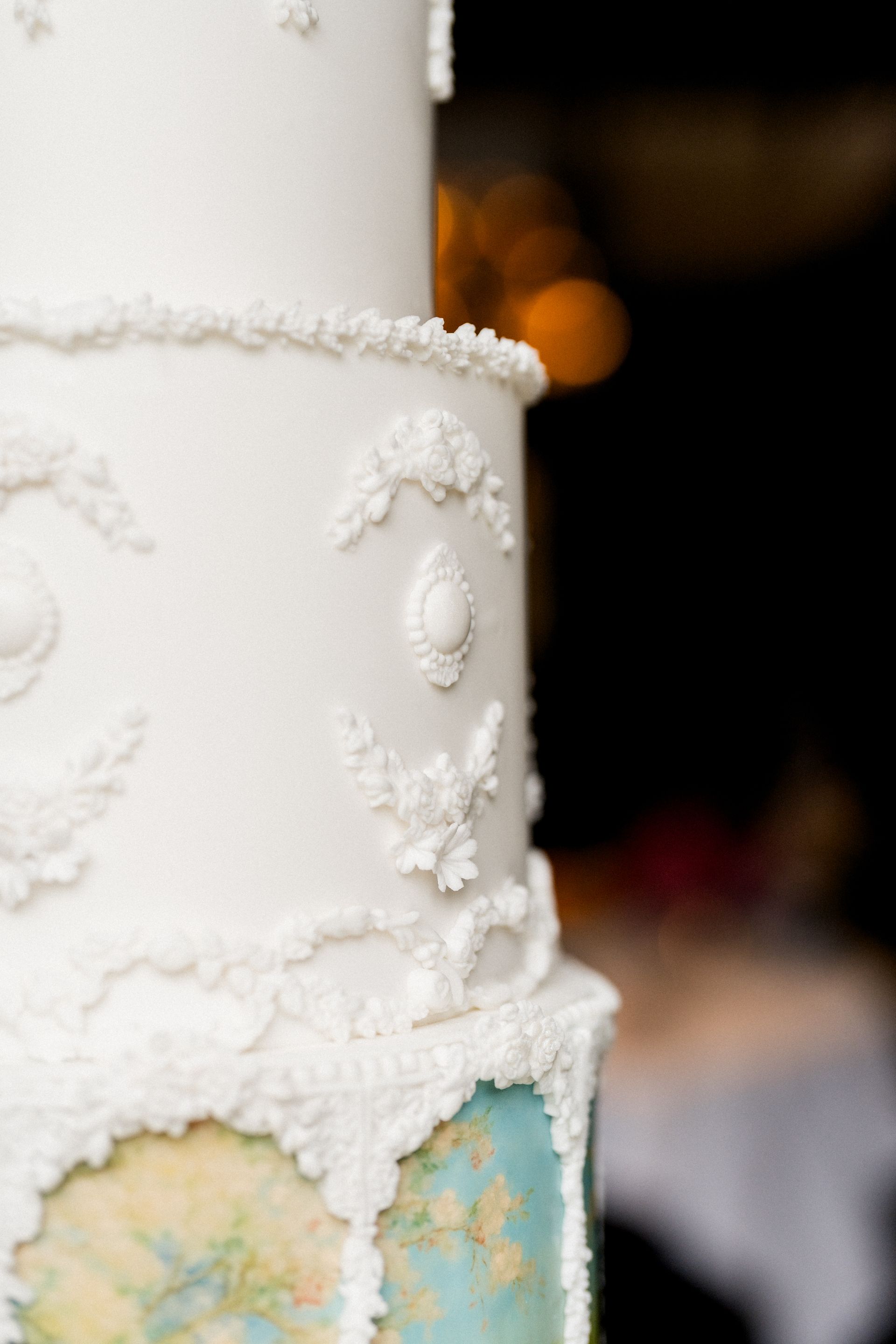 Close-up van de bovenste etages van de witte bruidstaart, met handgemaakte witte suiker ornamenten: festoenen, bloemmotieven en een ovaal medaillon in klassieke stijl.