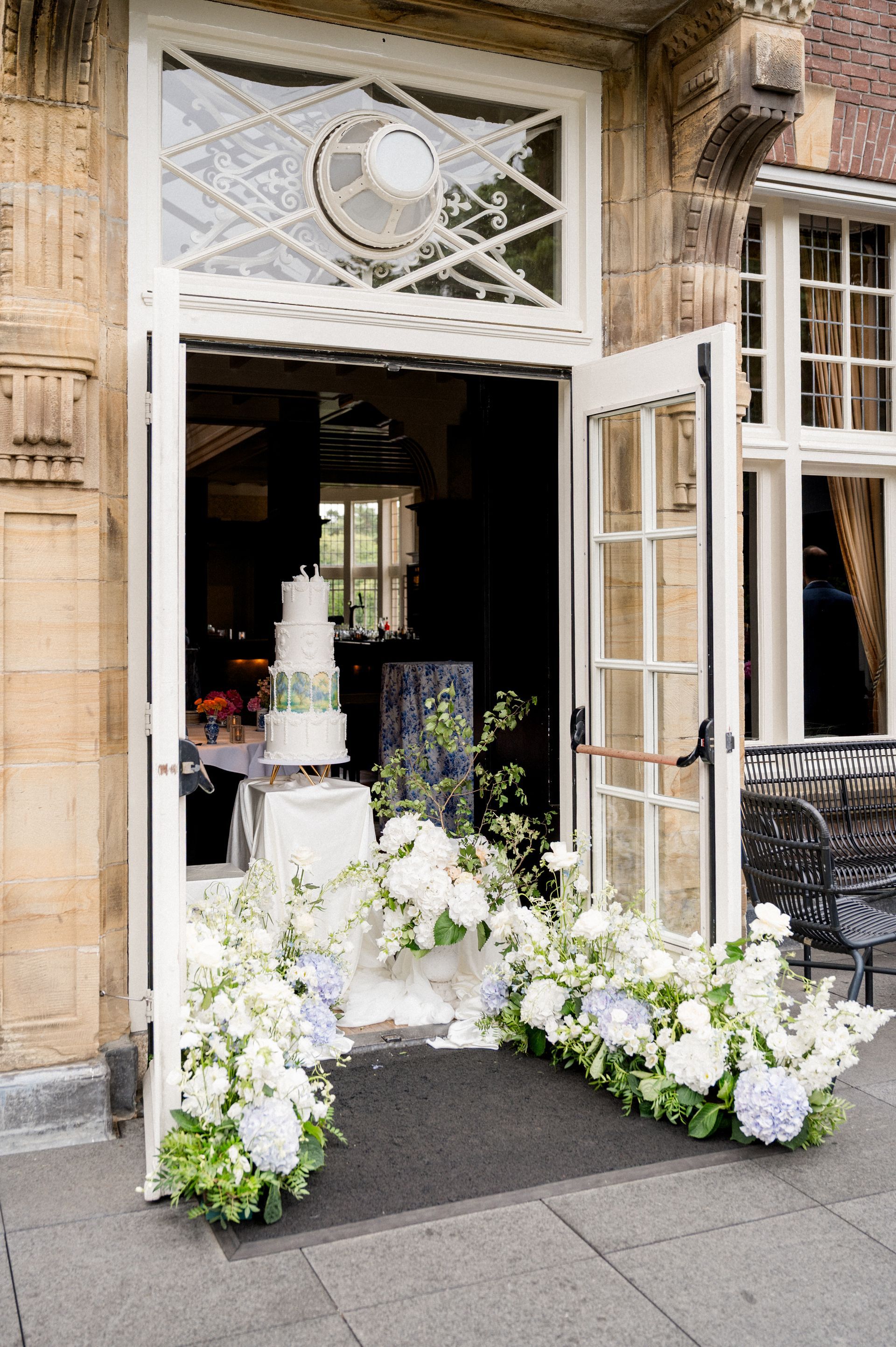 Vierlaagse witte bruidstaart op gouden standaard, opgesteld in de openstaande kasteeldeuren van Kasteel de Hooge Vuursche, omringd door een weelderige bloemenzee van witte hortensia's en ranke groene takken. 