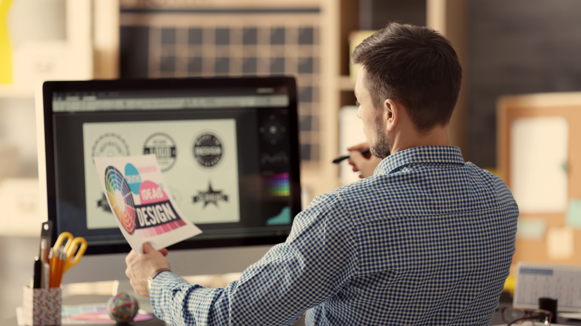Graphic designer, at a desk, looks at design on paper, while working on a computer.