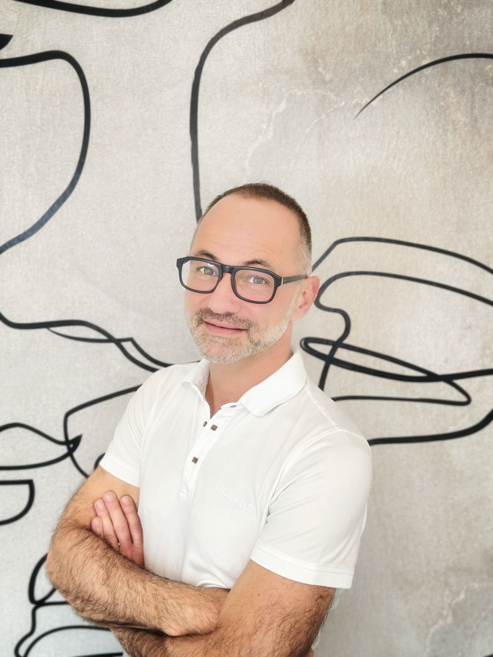 Ein Mann mit Brille und weißem Hemd steht vor der Zeichnung eines Gesichts an einer Wand.