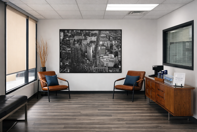 Modern office waiting room with two orange chairs, a wooden cabinet, and a black-and-white city artwork.