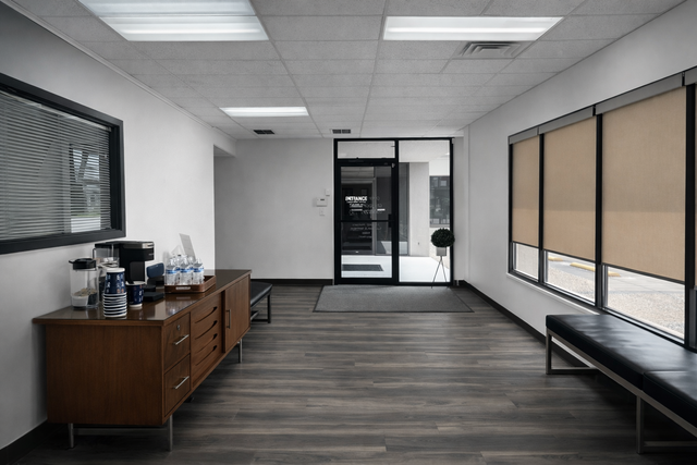 Empty office waiting area with wood cabinet, bench, and large windows on gray carpet.
