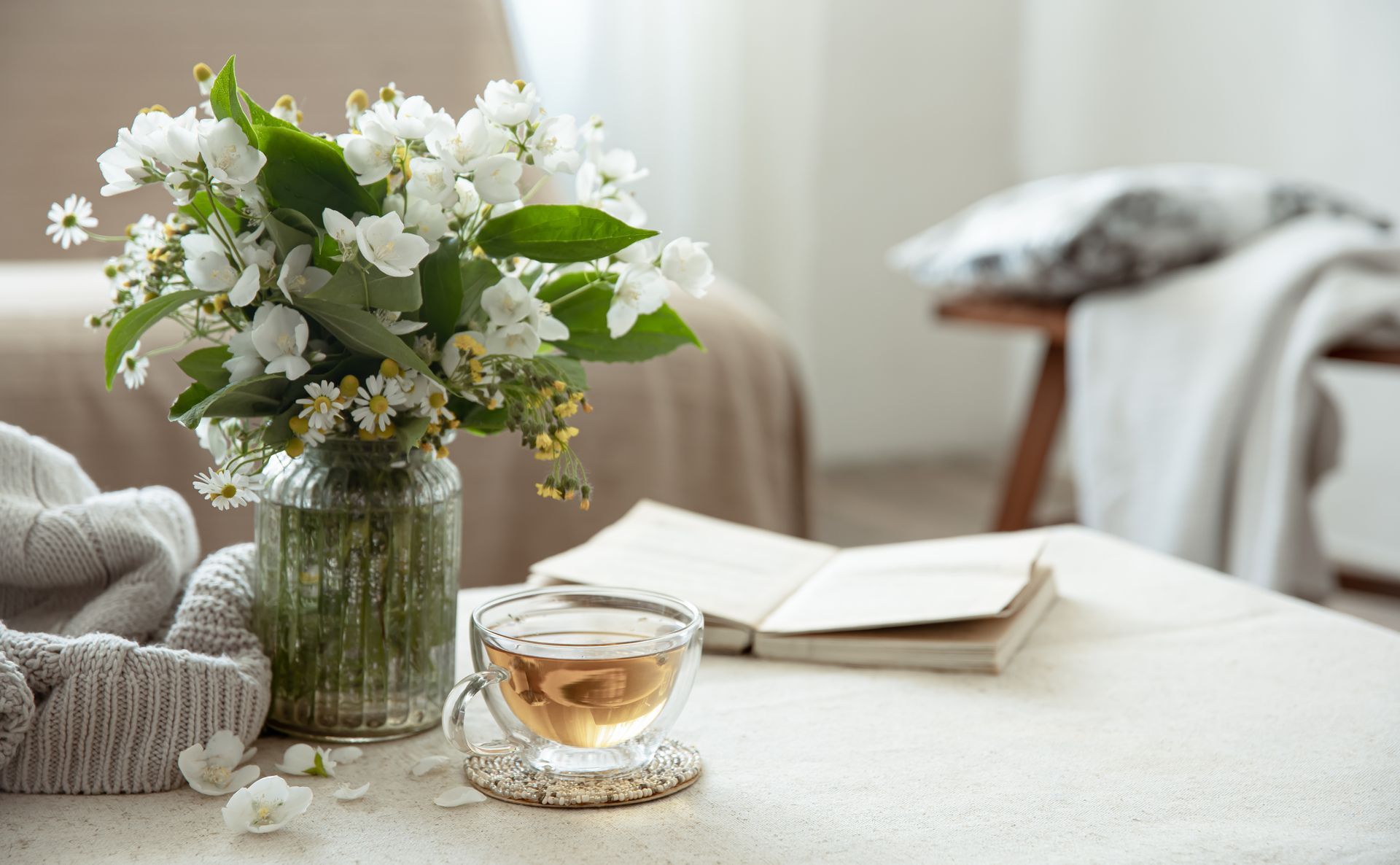 Vase of white flowers, cup of tea, open book, and cozy blanket on a table in a bright room.