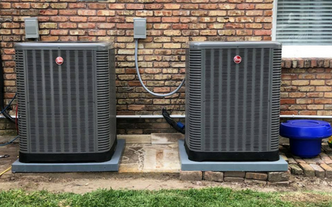 Two Rheem air conditioning units sit on grey pads against a brick exterior wall.