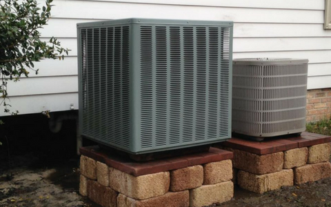 Two gray residential air conditioning units sit on raised brick platforms against a white horizontal-siding wall.