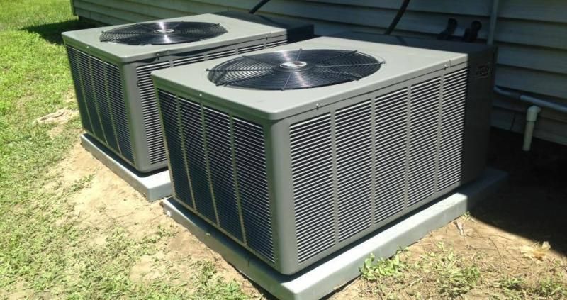 Two grey central air conditioning units sit on concrete pads beside the exterior wall of a building near a grass lawn.
