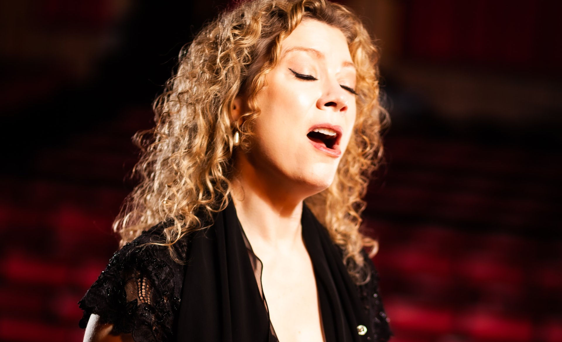 A woman with curly hair is singing into a microphone in a dark room.