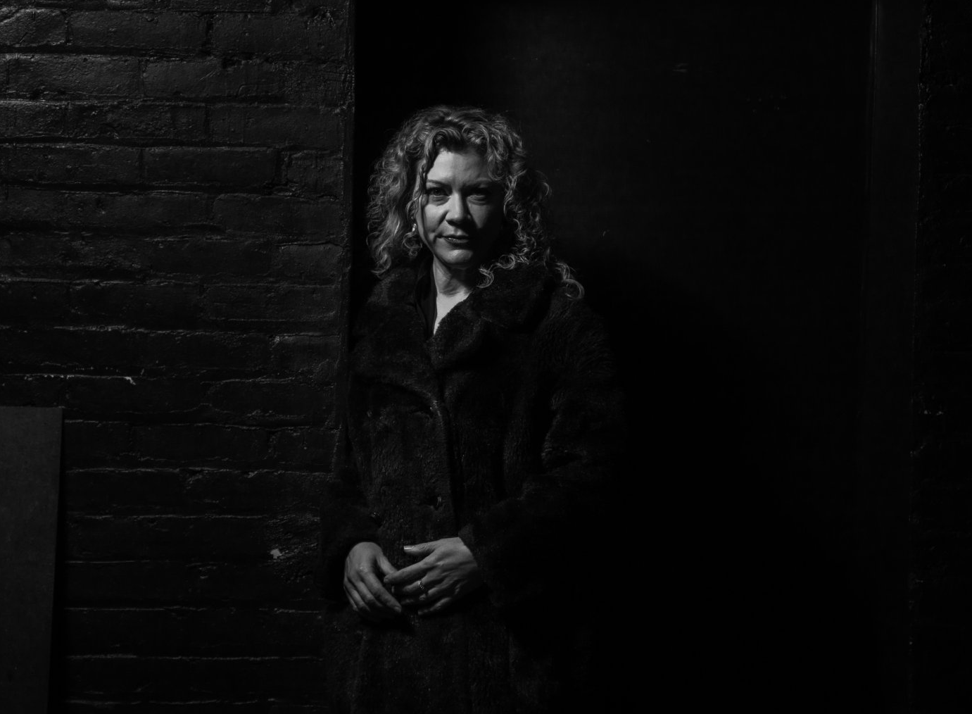 A black and white photo of a woman in a fur coat standing next to a brick wall.
