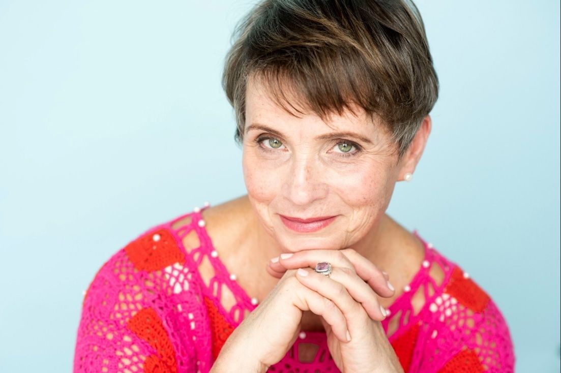 A woman in a pink shirt is sitting with her hands folded and smiling.