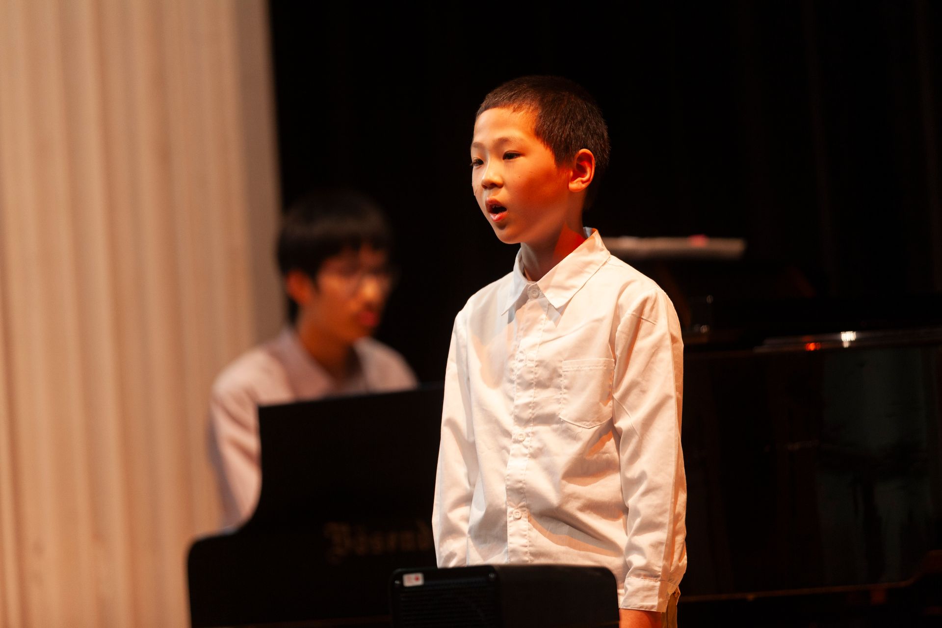 A young boy is singing on stage in front of a piano.
