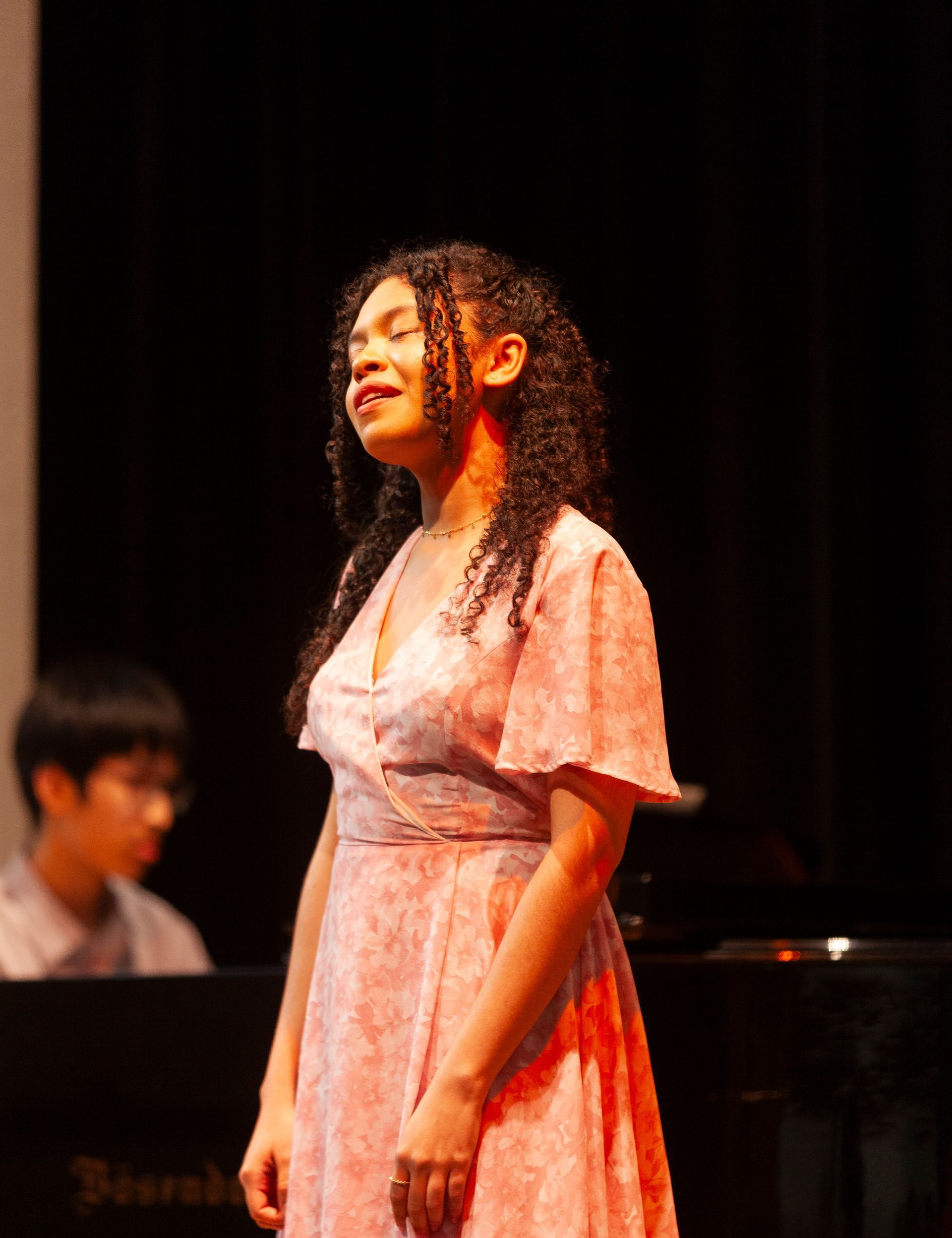 A woman in a pink dress is singing on a stage in front of a piano.